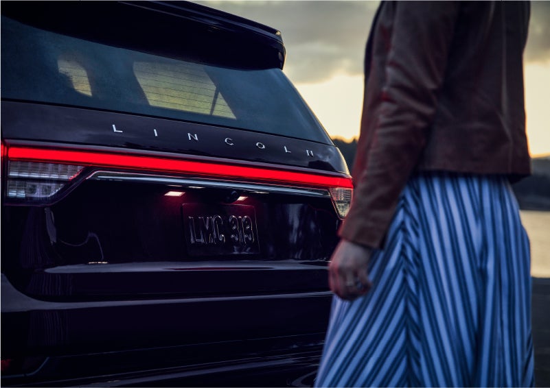 A person is shown near the rear of a 2023 Lincoln Aviator® SUV as the Lincoln Embrace illuminates the rear lights | John Vance Auto Group in Guthrie OK
