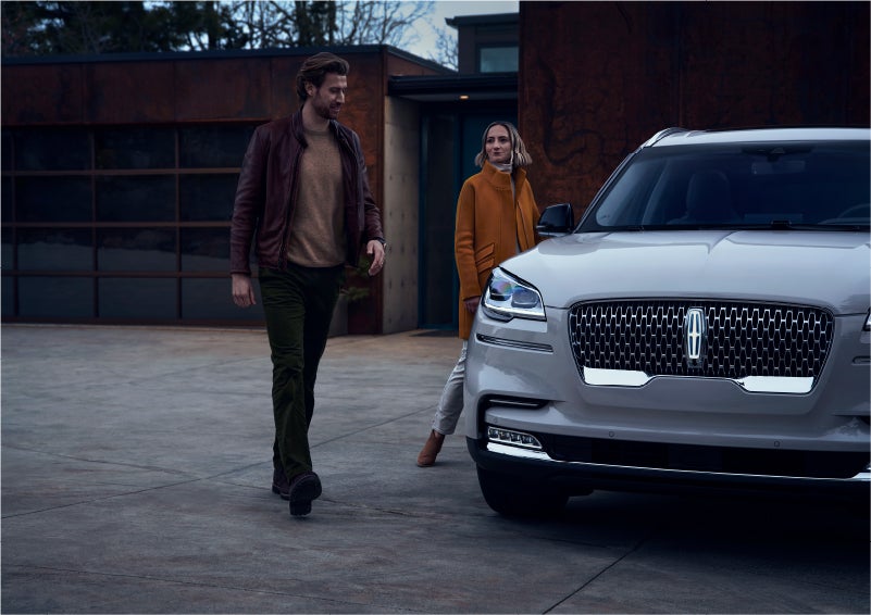 A man and a woman approaching a 2023 Lincoln Aviator® SUV, which illuminates certain lights when they are close | John Vance Auto Group in Guthrie OK
